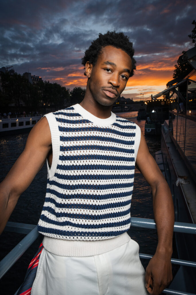 Portrait of a man in a white-and-navy striped knit vest standing on a railing by water at sunset.