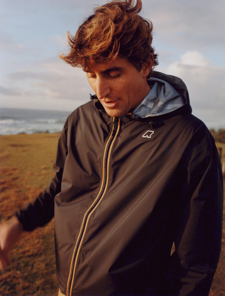 Young man with tousled brown hair wearing a dark jacket with a light hood of capsule collaboration between K-Way x Leonardo Fioravanti, looking down near a windy beach.