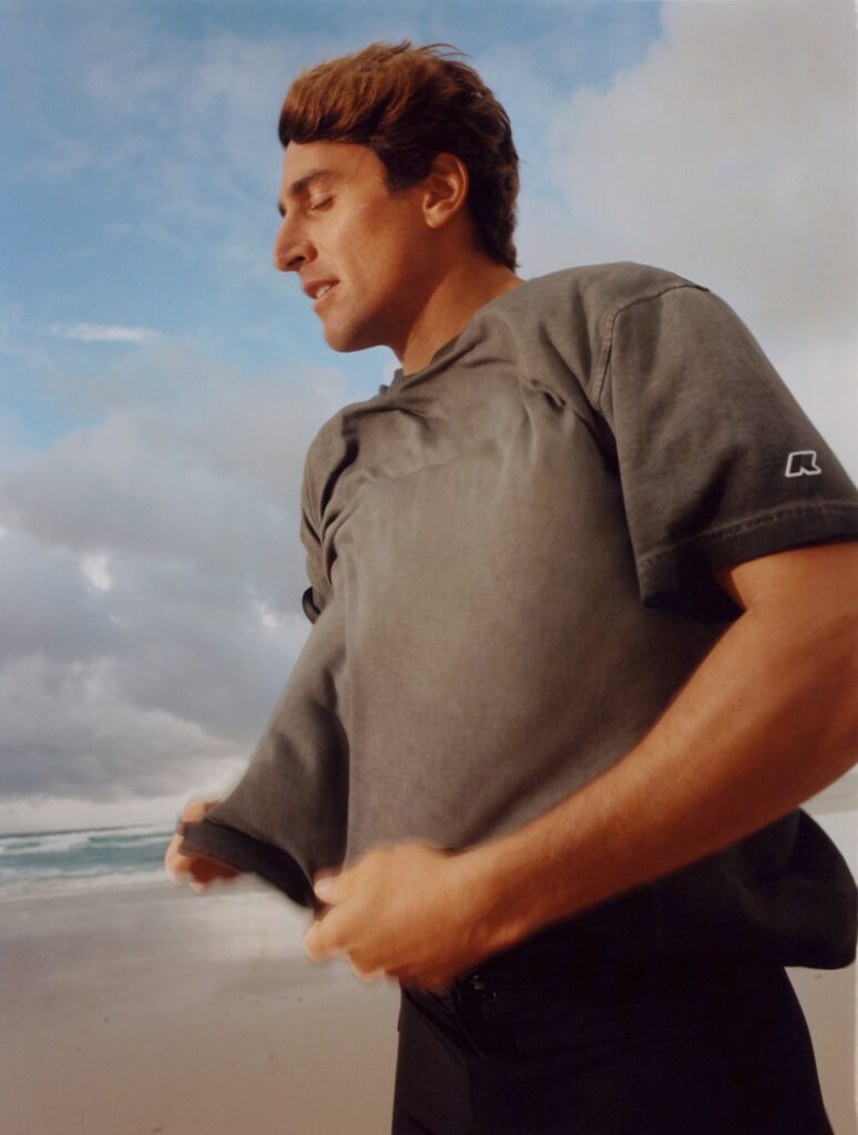 Side-profile of a man in a gray t-shirt from capsule collaboration between K-Way x Leonardo Fioravanti standing with arms crossed on a beach, cloudy blue sky above.
