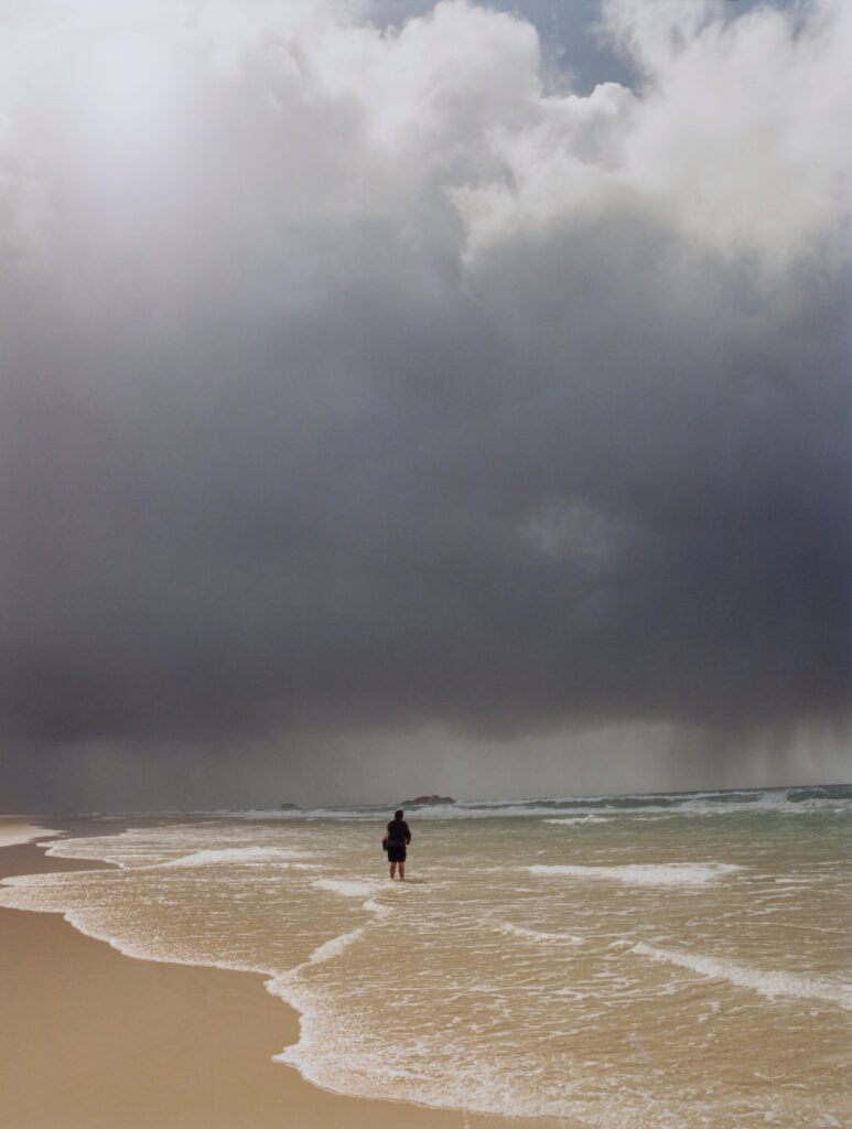 A lone person stands at the edge of a sandy beach, facing a vast, dark stormy sky over rough ocean waters as a massive rain cloud approaches.