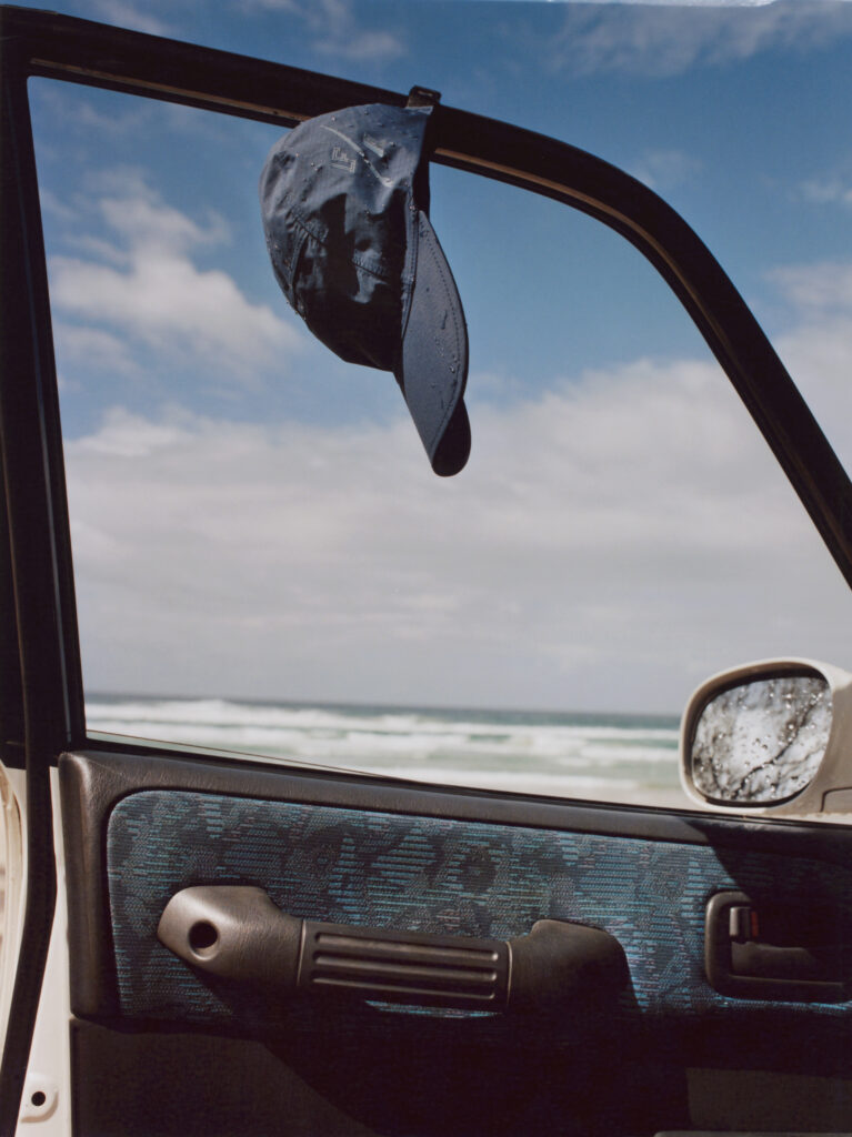 Blue cap hanging from a car window frame against a clear seaside sky, with ocean waves in the background.