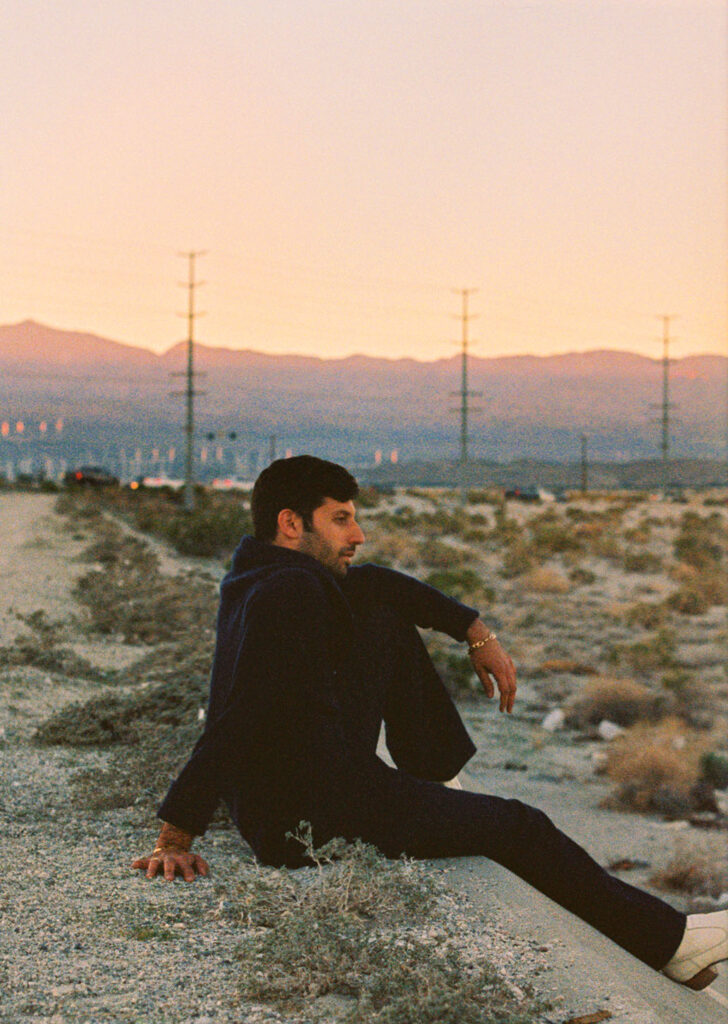 Joseph Reuben sits on a rocky desert slope at sunset, facing right, with distant power lines and mountains on the horizon.