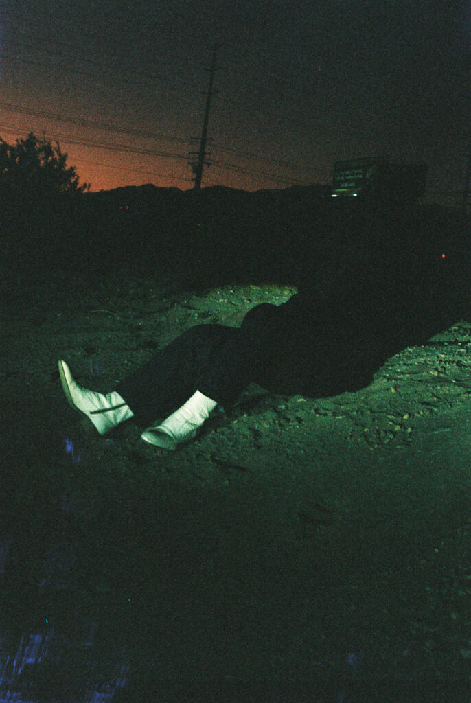 Dark outdoor night scene with Joseph Reuben lying or seated on rocky ground, white boots visible, silhouettes of trees and a distant illuminated sign on the right.