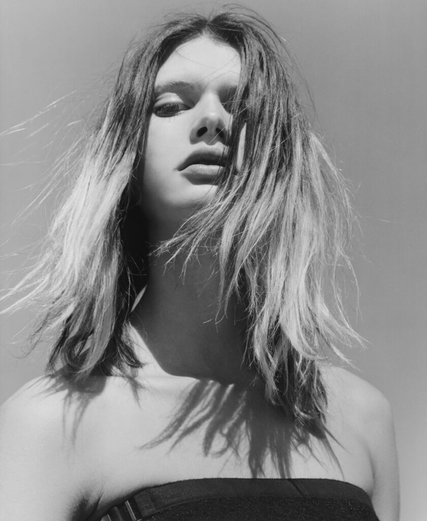 Black-and-white close-up portrait of a young woman with wind-swept hair across her face against a clear sky.