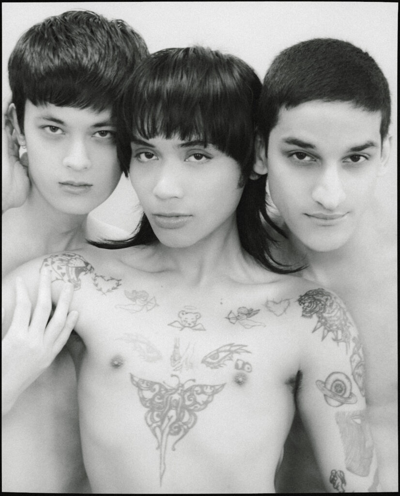Three shirtless young adults posing closely; two women on the left and a man on the right, with tattoos on the chest and arms visible.