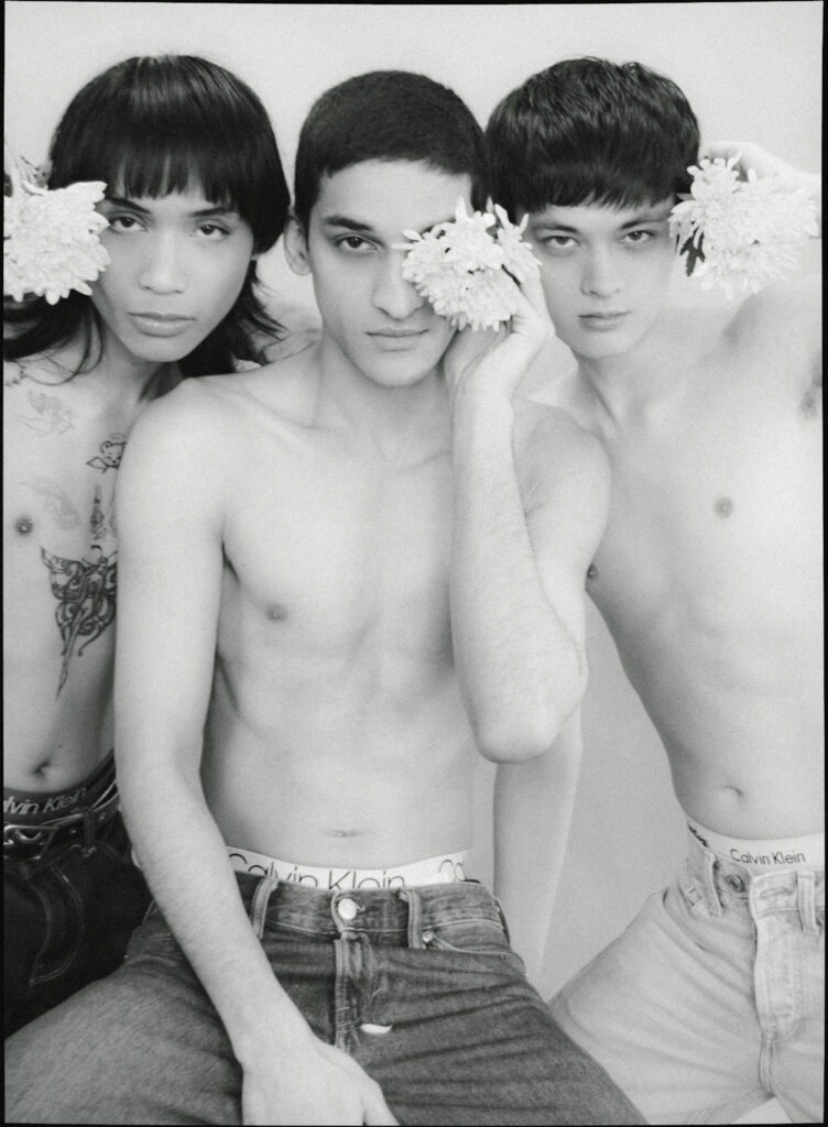 Three shirtless young men posing closely, each holding or touching white flowers near their faces in a black-and-white photo.
