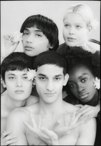 Black-and-white group portrait of six young people with flowers in their hair, posed closely together.
