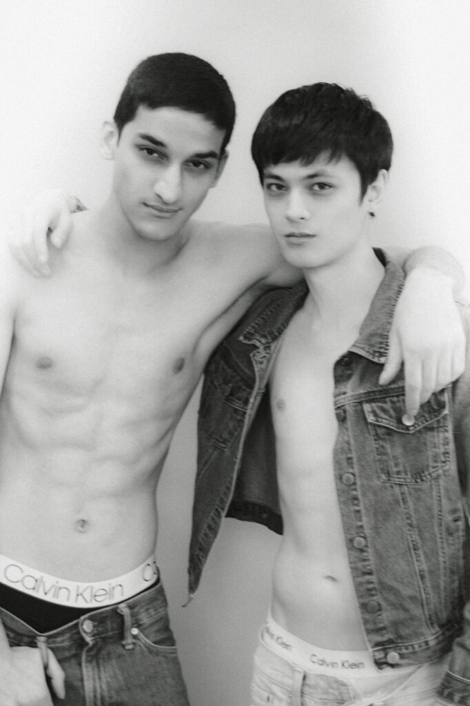 Two young men pose with arms around each other; one shirtless in jeans and Calvin Klein underwear, the other in a denim jacket.