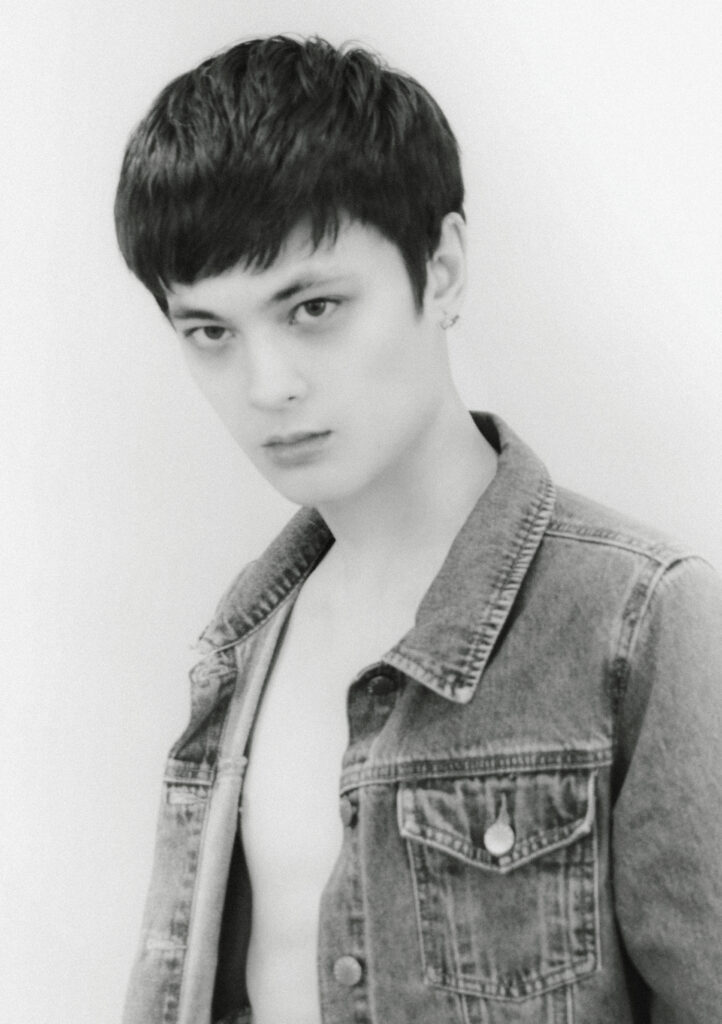 Black-and-white portrait of a person with short dark hair wearing a denim jacket, looking slightly to the left.
