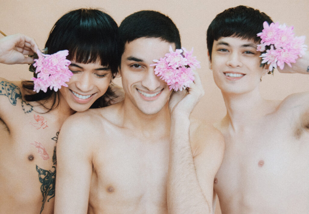 Three topless friends smile, each holding pink chrysanthemum flowers to their cheeks. A light, warm-toned portrait.