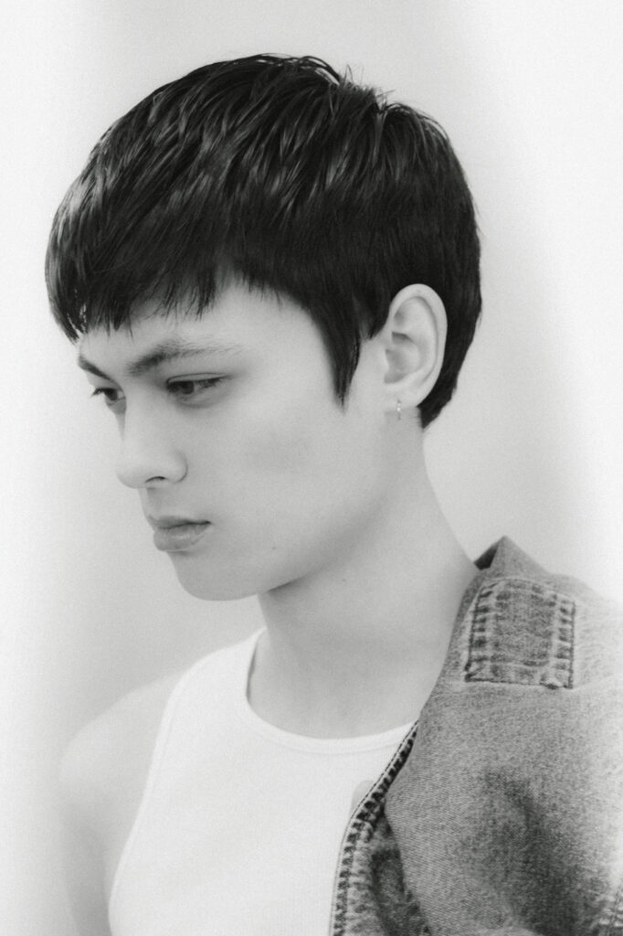 Close-up side profile of a young person with short dark hair, wearing a white tank top and a denim jacket over the shoulder, looking down.