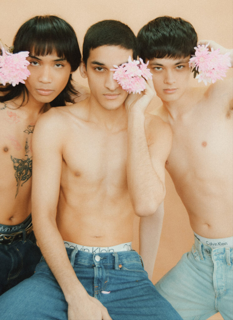Three topless young men pose against a peach background, each holding pink flowers near their faces; tattoos visible on the left arm, wearing jeans and Calvin Klein underwear.