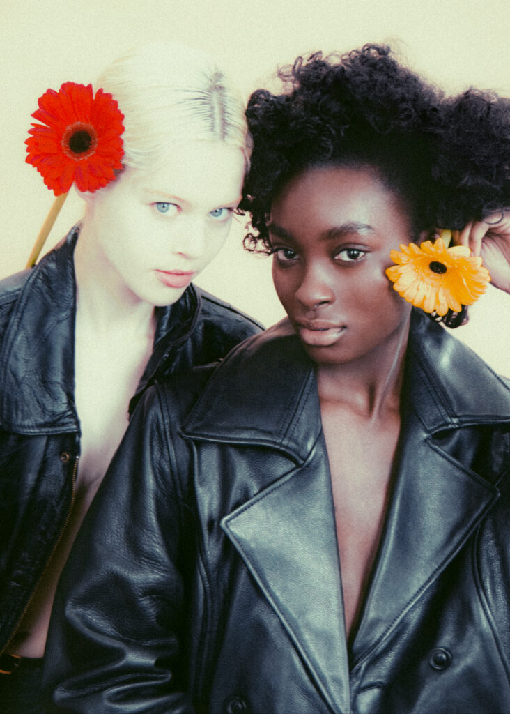 Two women in black leather jackets pose close together, each with a bright flower behind the ear—a red gerbera on the left and a yellow one on the right.