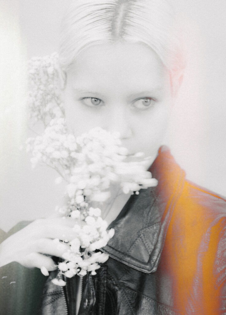 Person holding a white flower bouquet close to their face, with only the eyes visible and wearing a denim jacket. Orange sleeve shows on the right.
