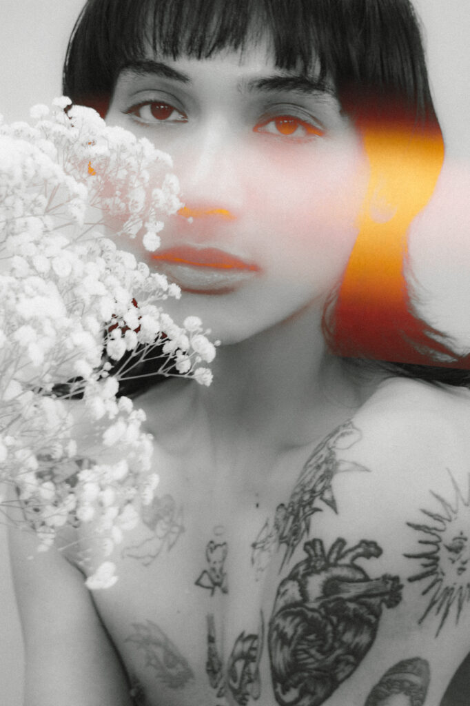 Close-up portrait of a woman with a bouquet of white flowers near her face, grayscale with orange accents on lips and one eye and a tattooed chest visible.