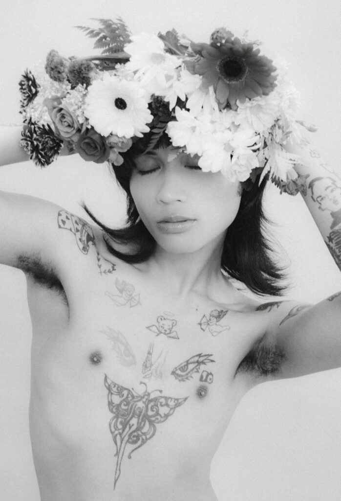Topless person wearing a large flower crown with eyes closed; visible tattoos on arms and chest in black-and-white portrait.