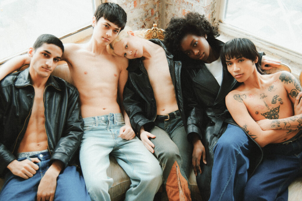 Group of five young people lounging together on a couch, some shirtless, wearing jeans and leather jackets in a casual setting.