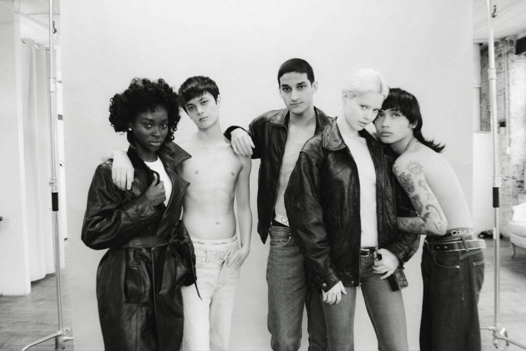 Five young models pose together in a studio wearing leather jackets; two are shirtless, showing jeans and accessories, in black-and-white.