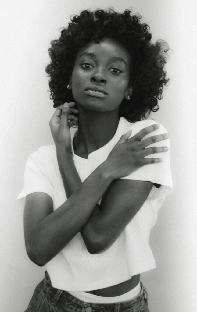 Black woman with curly hair wearing a white t-shirt and jeans, posing with arms crossed and hand touching neck.