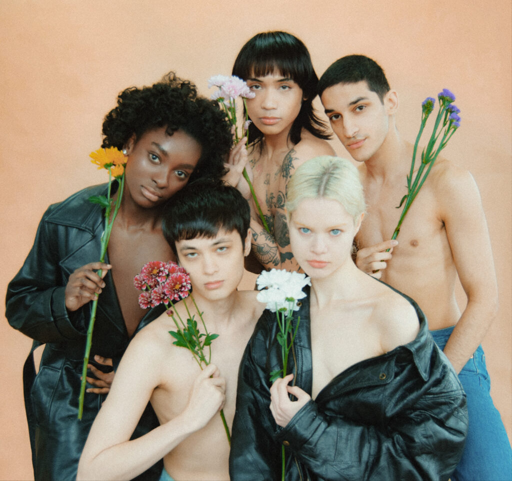 Group of diverse young adults posing together with flowers against a peach backdrop; topless with denim jackets, casual and intimate portrait composition.