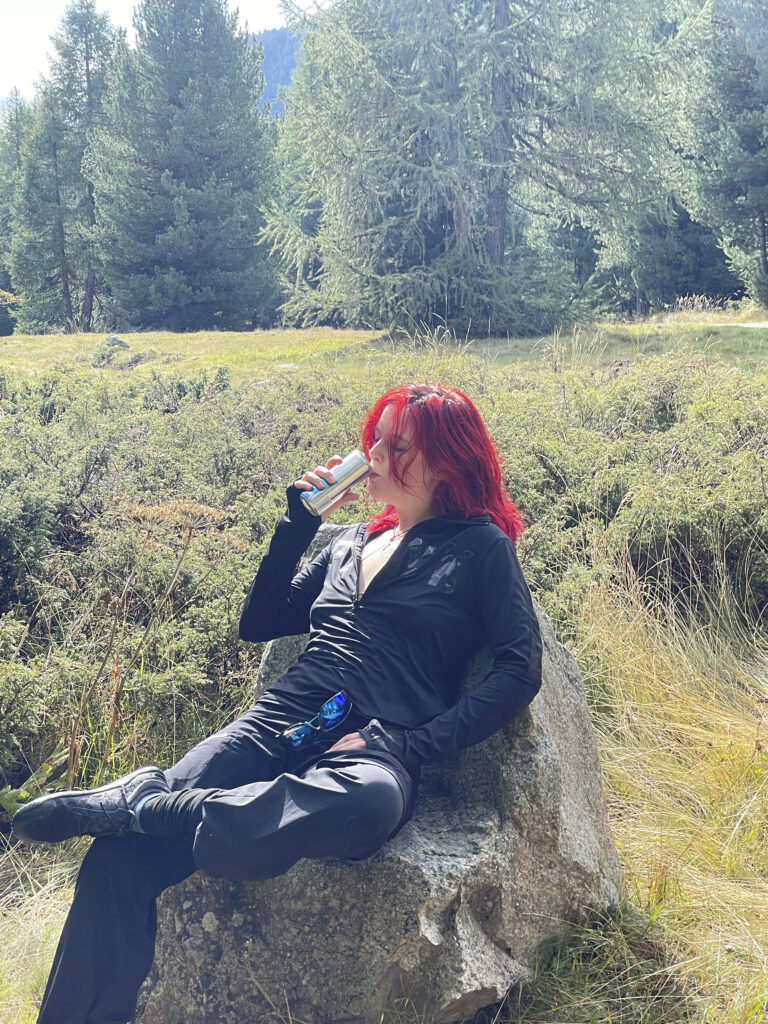 Person with bright red hair in a black outfit sits on a large rock in a forest clearing, drinking from a metal bottle/ flask.