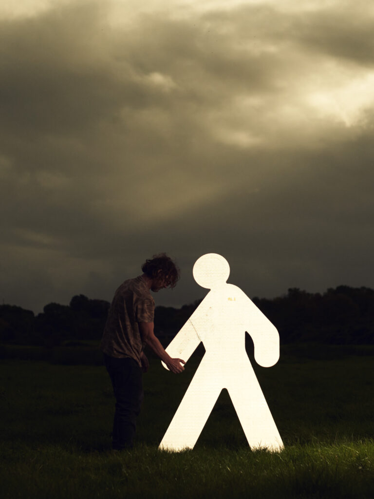 Man standing beside a large lit white pedestrian figure sign in an open field at dusk.