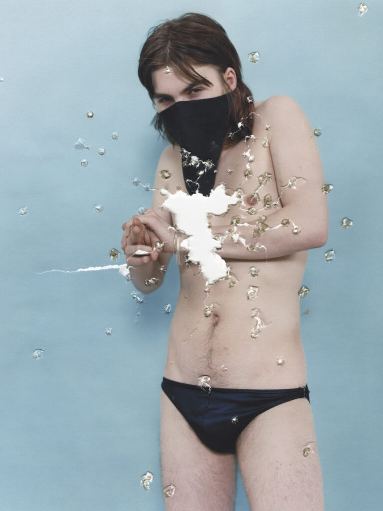 Young adult wearing a black face mask and dark underwear against a blue backdrop, with torn white patches scattered across the image.