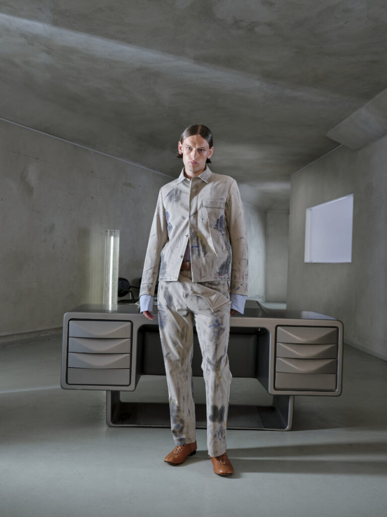 Silhouette A.A. Spectrum SS27 en veste blanche texturée et pantalon gris, dans un décor brut et contemporain.