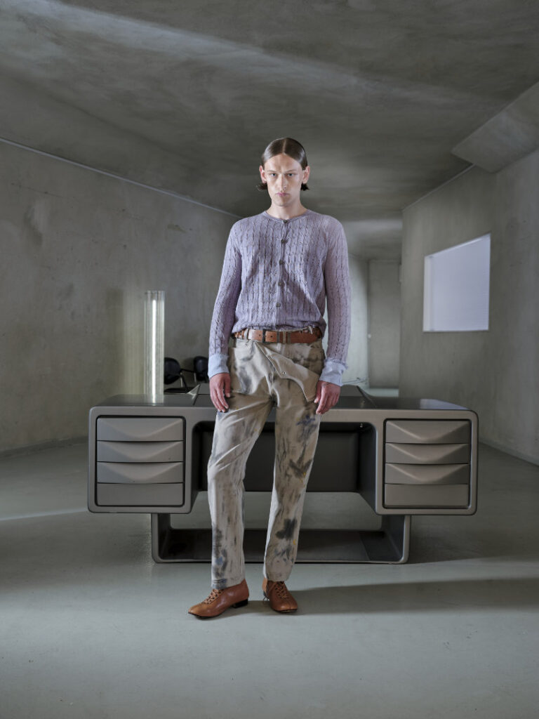 Person standing in a concrete, minimalist room behind a futuristic gray desk, wearing a lavender knit cardigan and paint-splattered tan pants with brown shoes.