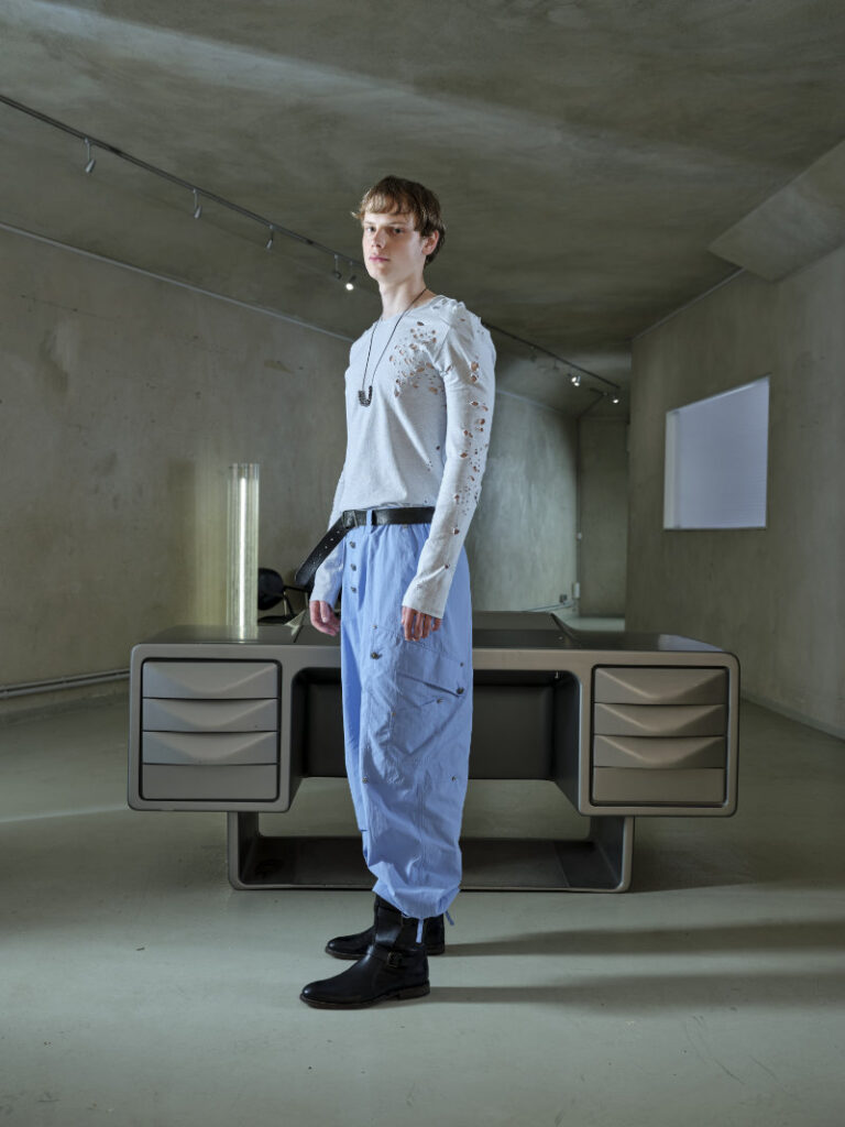 Model stands beside a futuristic desk in a gray studio, wearing a distressed white sweater and light blue pants, black boots.