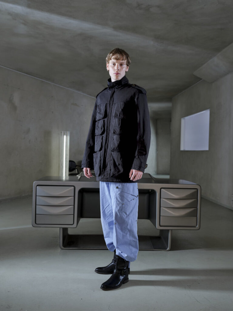 Model in a black utility jacket and light blue cargo pants stands in a bare concrete room beside a futuristic gray desk/console arcades behind him.