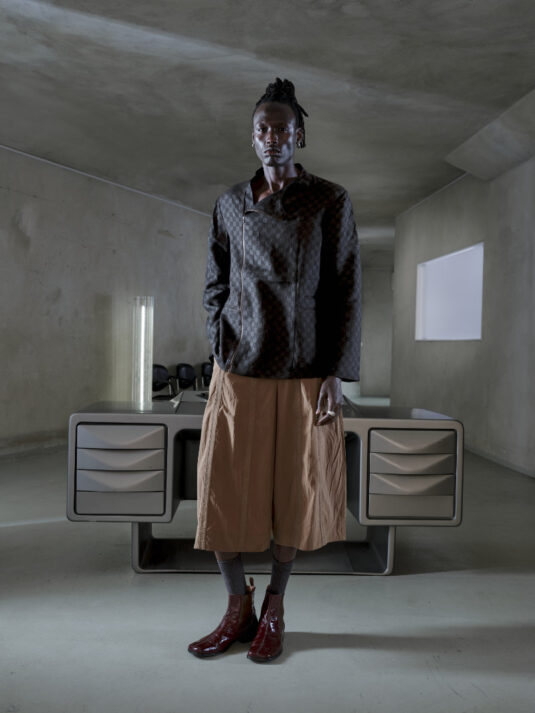 Male model in a textured dark jacket and wide tan shorts stands in a bare concrete room with a futuristic desk behind him.