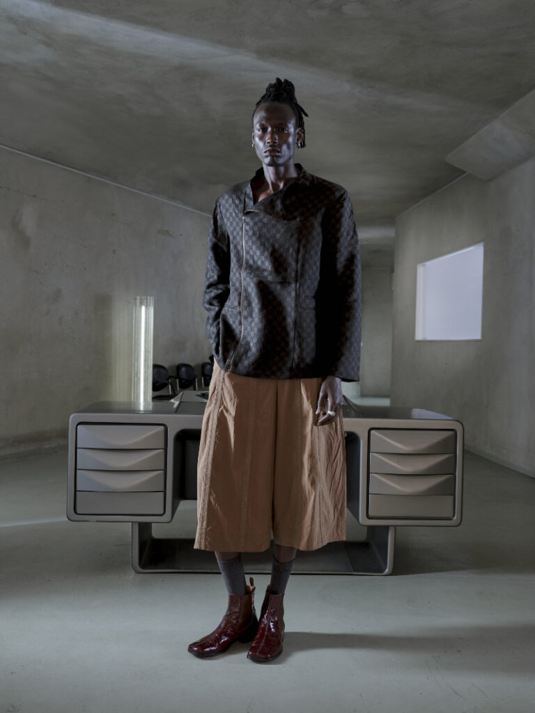 Male model in a textured dark jacket and wide tan shorts stands in a bare concrete room with a futuristic desk behind him.
