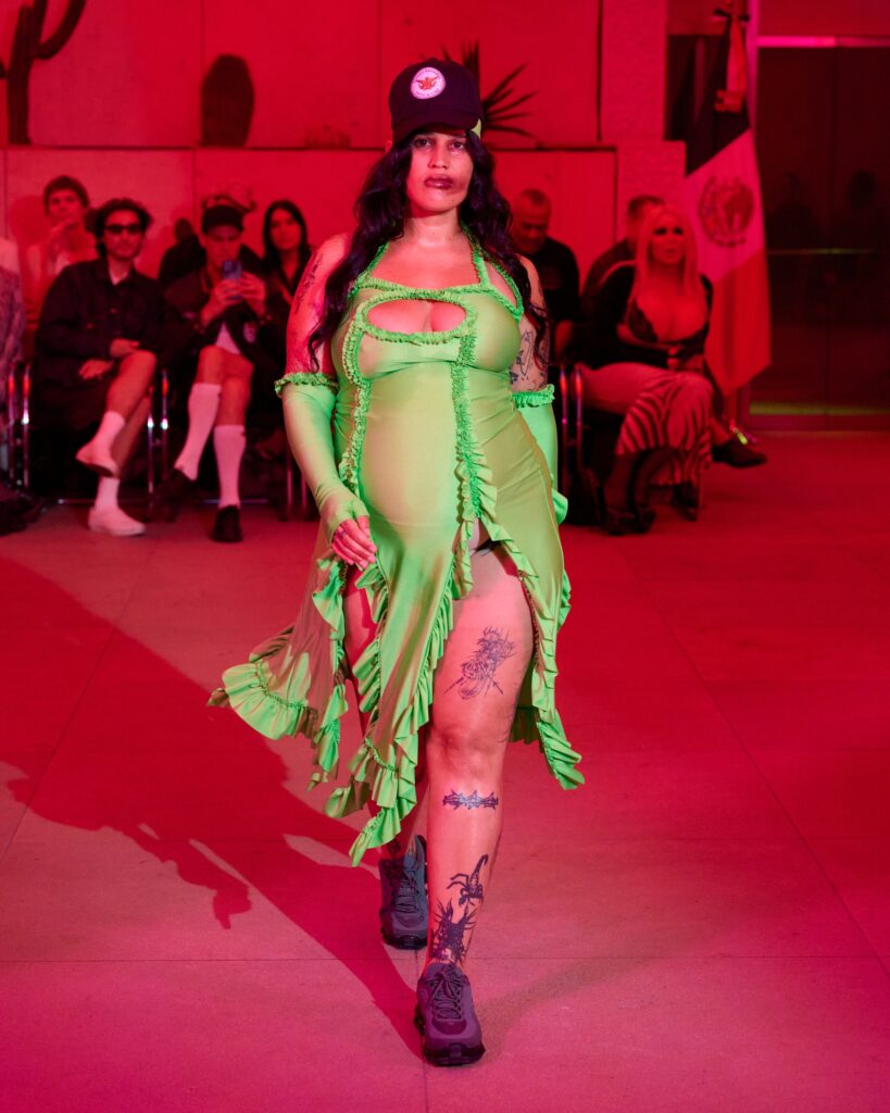 Model walking a runway in a bright green ruffled dress with cutouts, wearing a black cap; red-lit audience in the background.