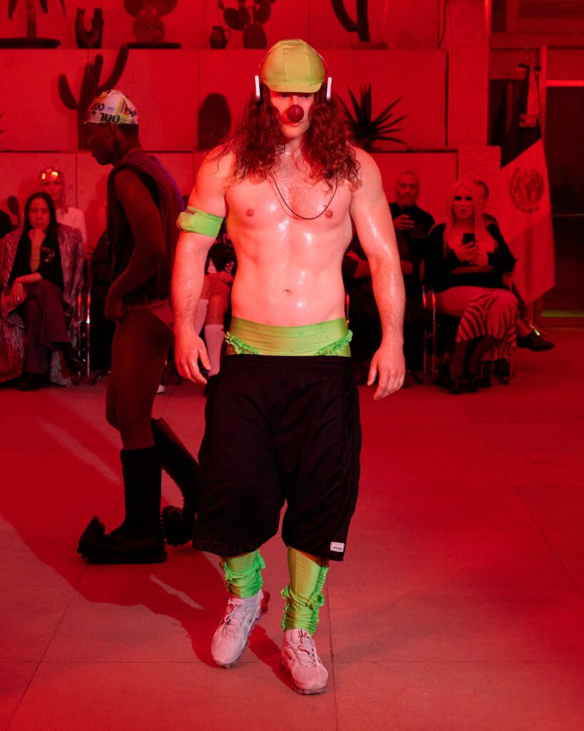 performer in neon green gear with a clown nose and long hair, skating on a red-lit stage while spectators watch in a dim venue