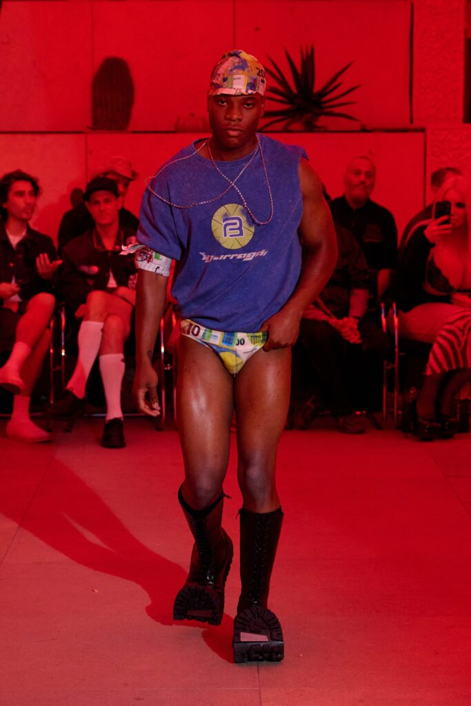 Male runway model wearing a blue sleeveless shirt, colorful underwear, and black boots under red lighting during a fashion show; audience seated in background