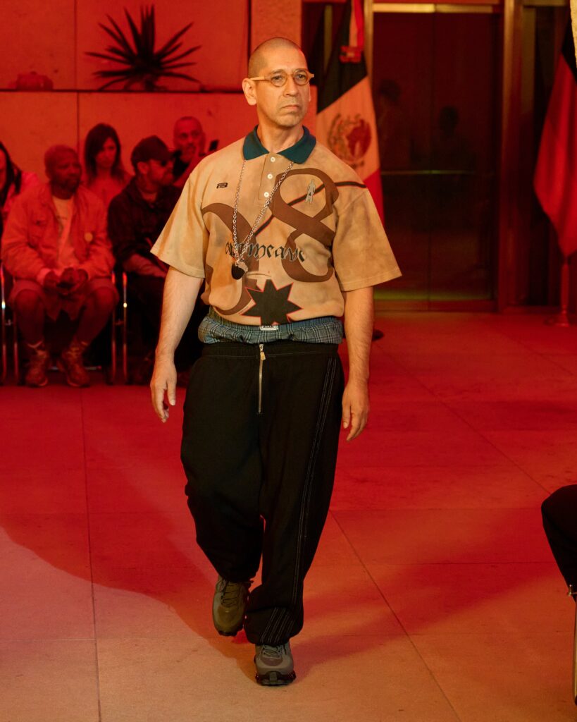Man with glasses and shaved head walks a runway in a tan graphic shirt and dark pants under red lighting; audience in background.