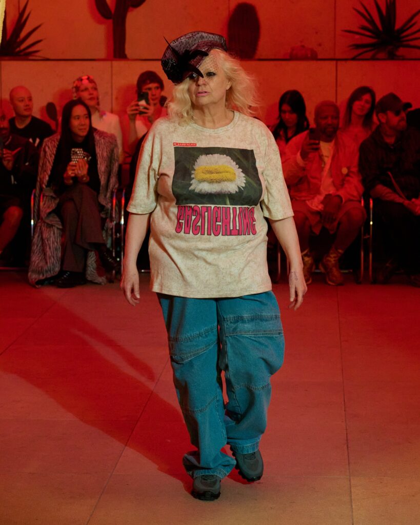 Model walking a runway in a beige graphic T-shirt with a daisy image, blue cargo pants, and a black feathered fascinator under red lighting, audience in background.