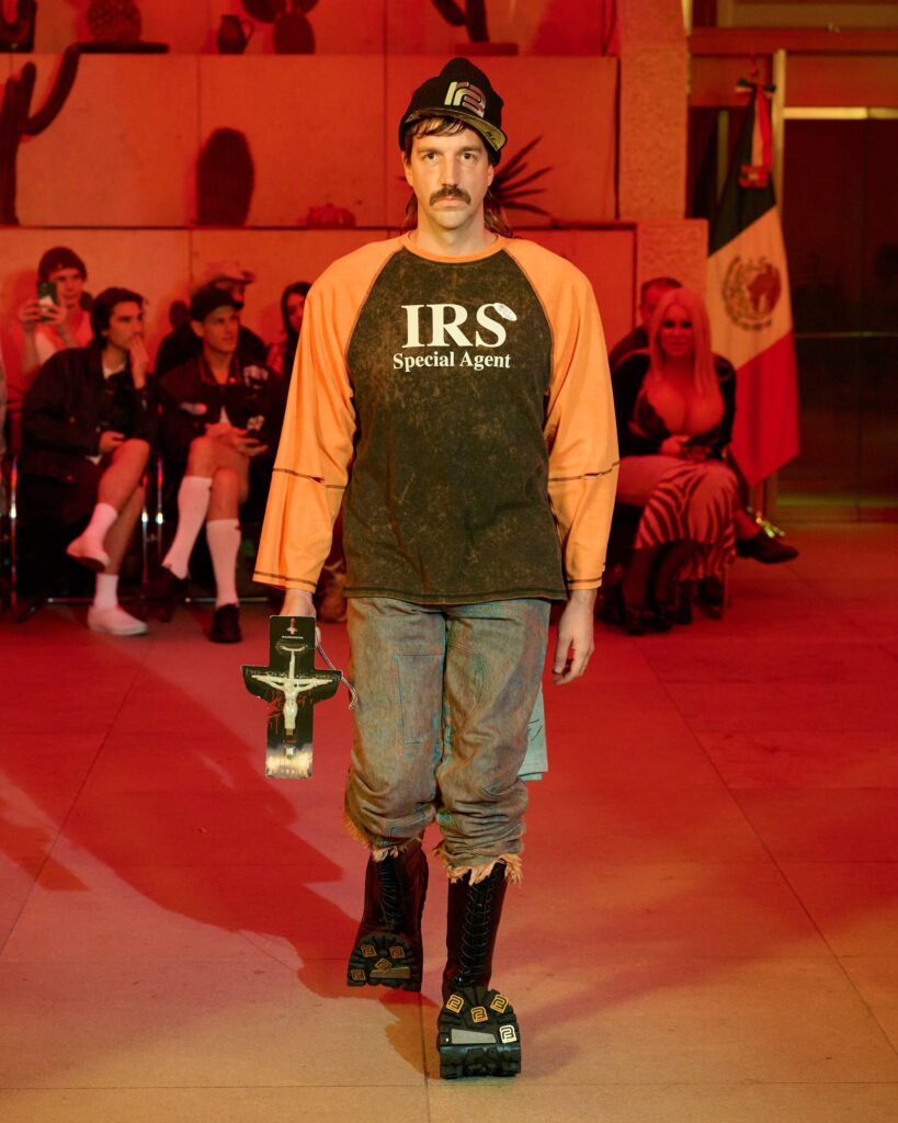 Male model with a mustache wearing an orange and black 'IRS Special Agent' shirt, walking on a runway under red lighting, audience in the background.