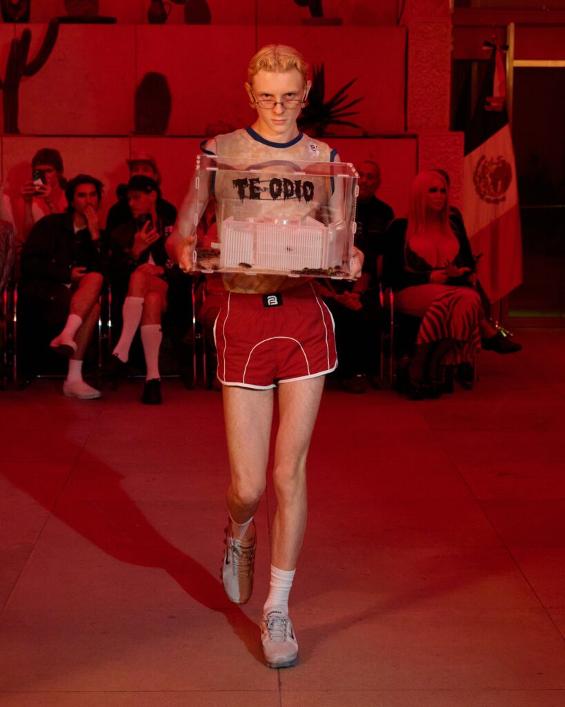 Model walking a runway while carrying a clear acrylic box with small turtles inside; box is labeled 'TE ODIO' under red lighting.