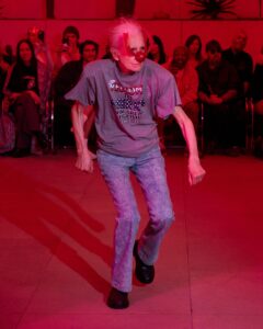 Older man wearing a red bird-shaped mask and a gray T-shirt dances in a red-lit room with an audience watching in the background.
