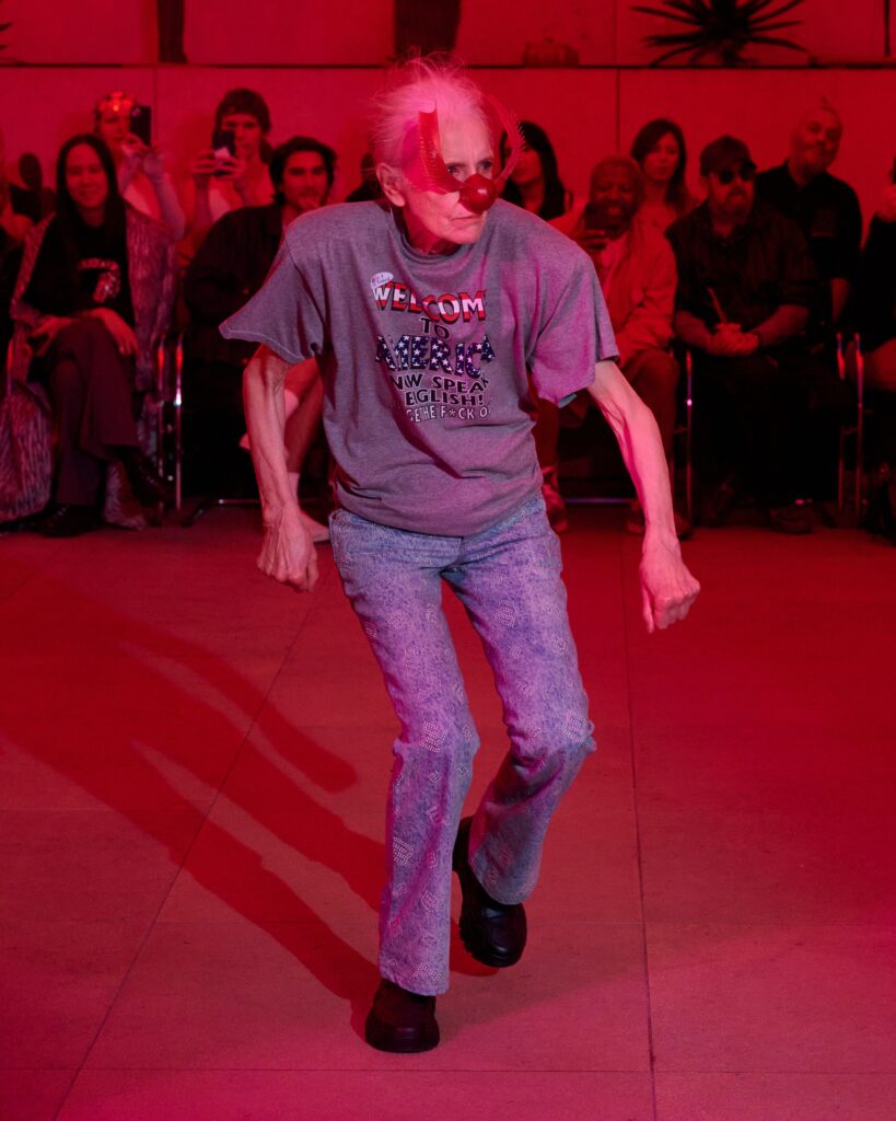 Older man wearing a red bird-shaped mask and a gray T-shirt dances in a red-lit room with an audience watching in the background.