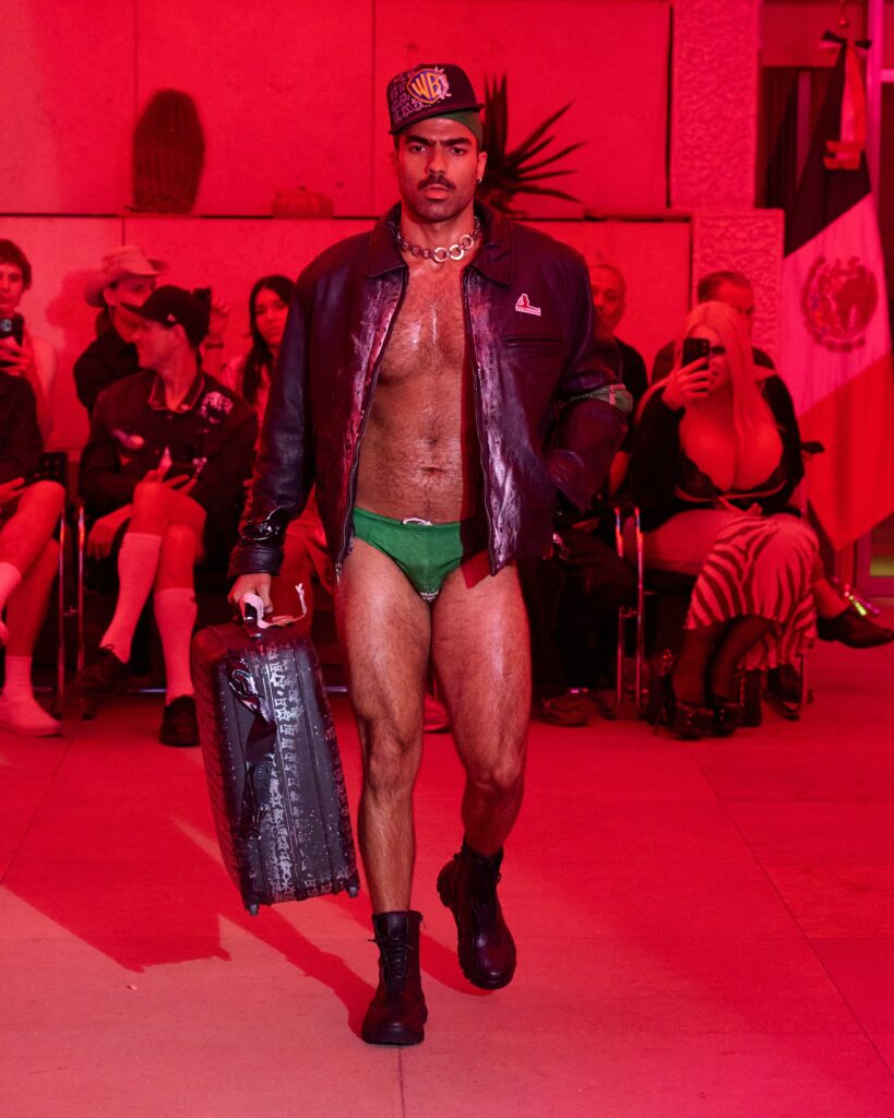 Male model walking the runway in an open black leather jacket, green briefs, cap, and chunky silver necklace under red lighting with spectators in the background.