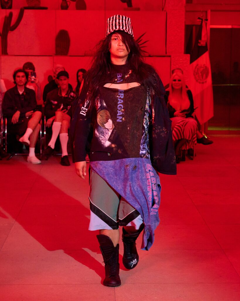 Model walking a fashion runway under red lighting, wearing a striped hat, long dark hair, distressed graphic shirt and layered skirt with tall black boots; audience seated in background.