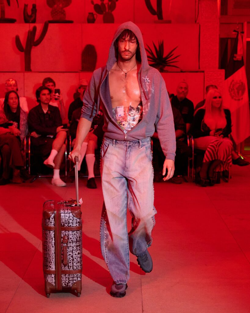 Male model on a runway in an open gray hoodie over a graphic shirt, pulling a patterned wheeled suitcase; audience seated in red lighting behind him.