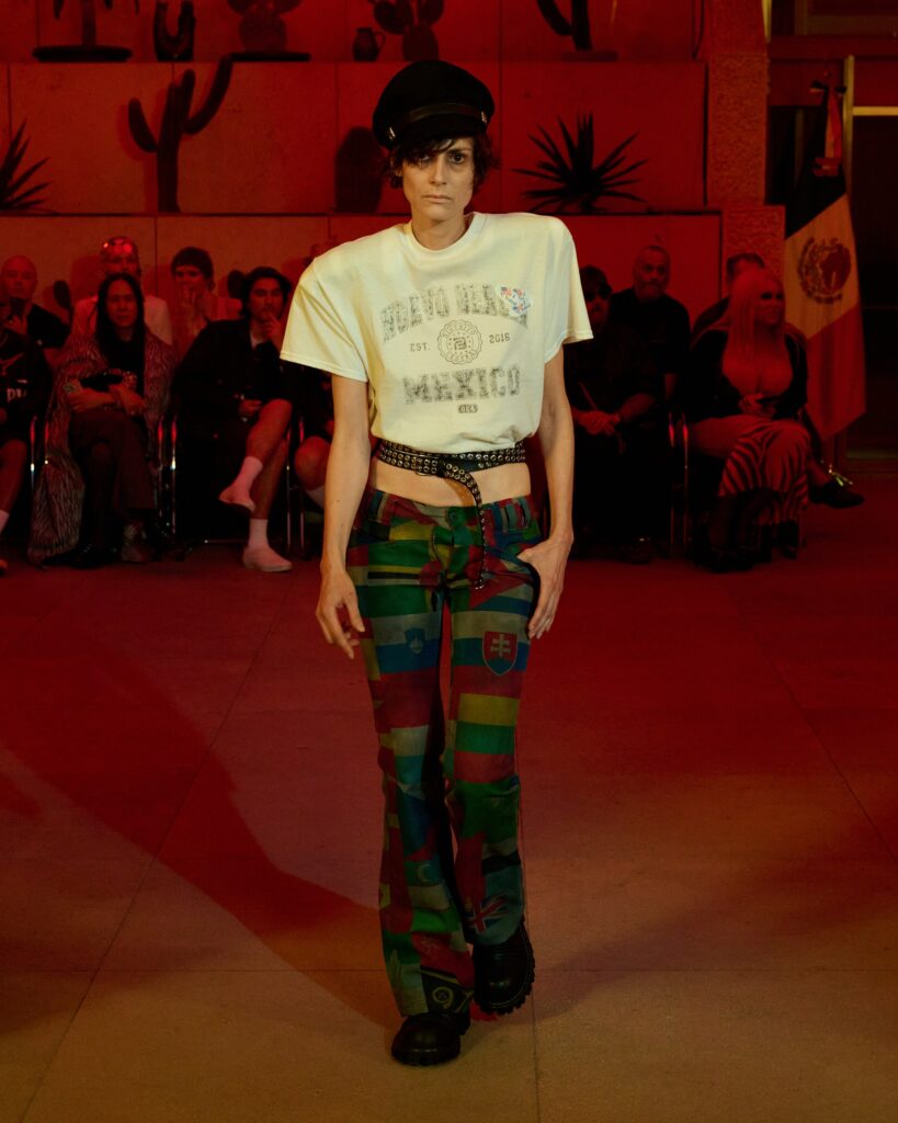 Model walks the runway in a white graphic tee, multicolor patchwork pants, a studded belt, and a black cap under red lighting.
