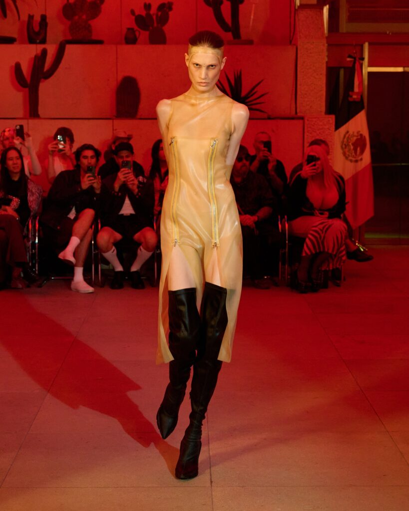 Model walks the runway in a sleeveless beige latex dress with visible zippers, paired with black knee-high boots under red lighting, audience in the background photographing the moment.
