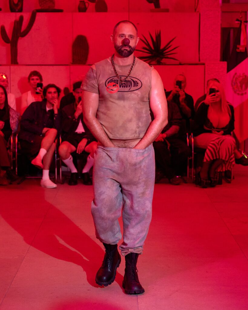 Male performer with a red clown nose walks a runway in a sleeveless tie‑dye shirt, chain necklace, and boots, under red lighting.
