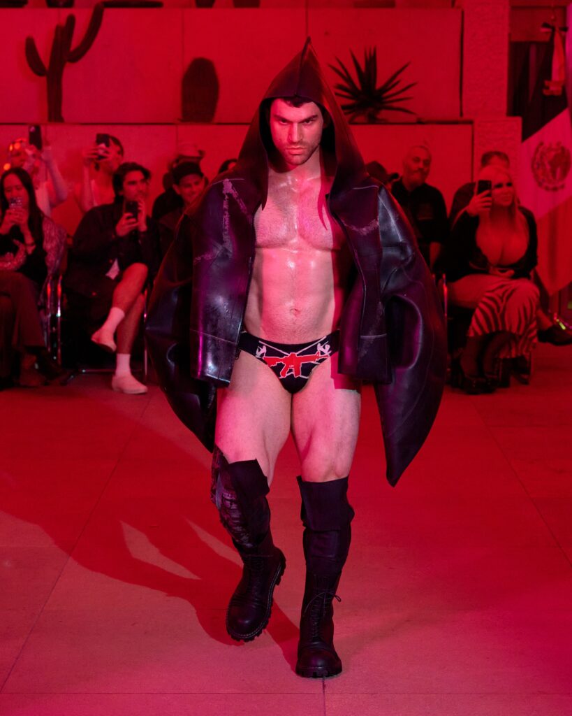 Male model walks the runway in a dark hooded cape and patterned red-and-black briefs, under red stage lighting with spectators in the background.