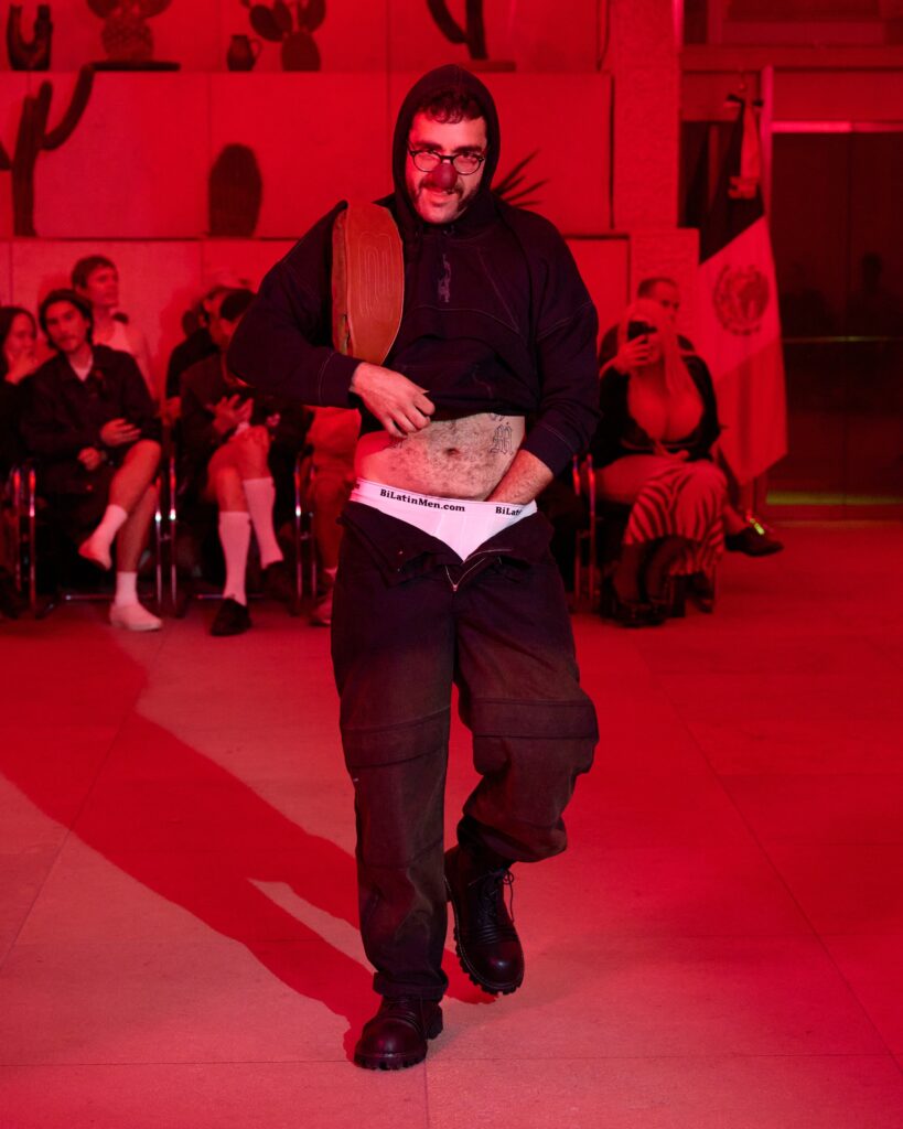 Man in black hoodie with a red clown nose walks on a red-lit runway, audience watching in the background.